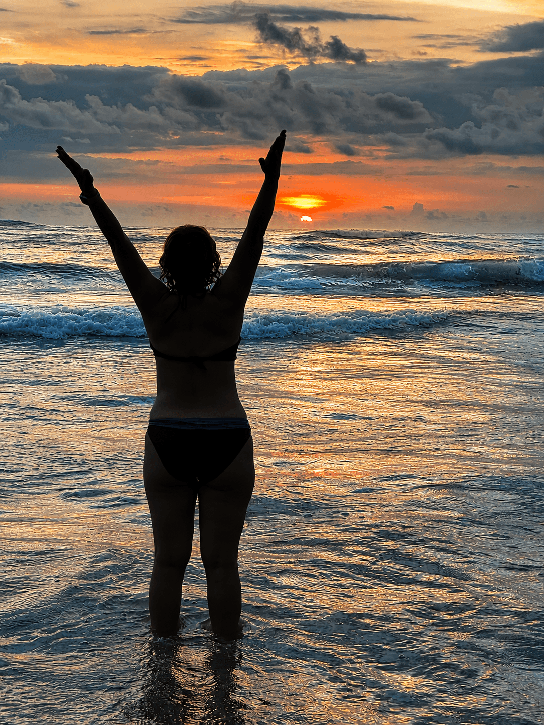 Woman standing in the ocean at sunset with arms raised